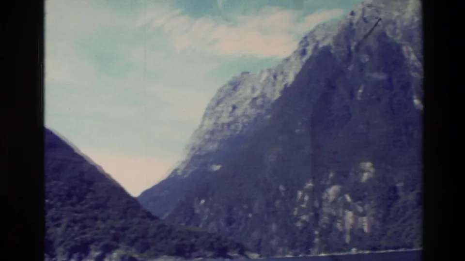 1982 New Zealand frame looking into a fjord-like gap between steep mountains with water and pale sky visible.