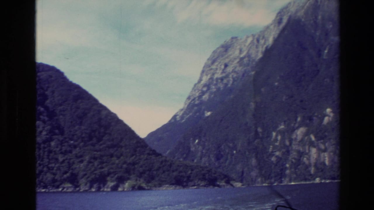 1982 New Zealand 8mm view from a cruise boat, showing dark fjord water and steep forested mountains under pale sky.