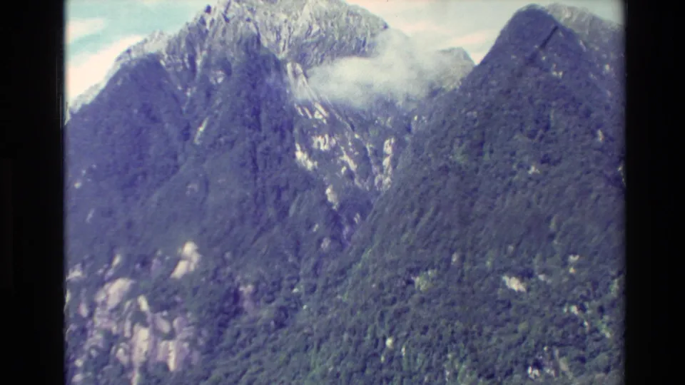 1982 New Zealand home-movie frame of a steep fjord mountain face, with forest cover, exposed rock, and low cloud.