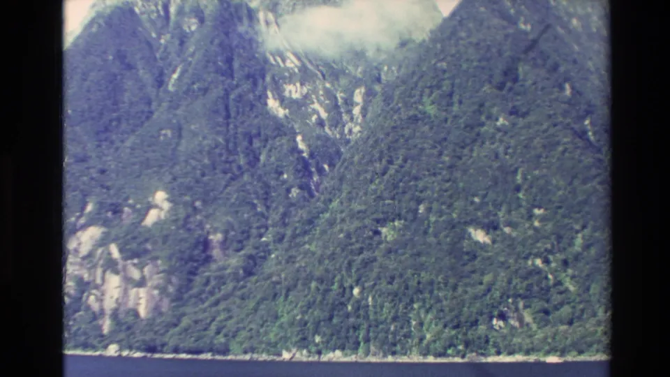 1982 New Zealand 8mm frame showing a forested fjord slope, scattered pale rock outcrops, and water at the base.