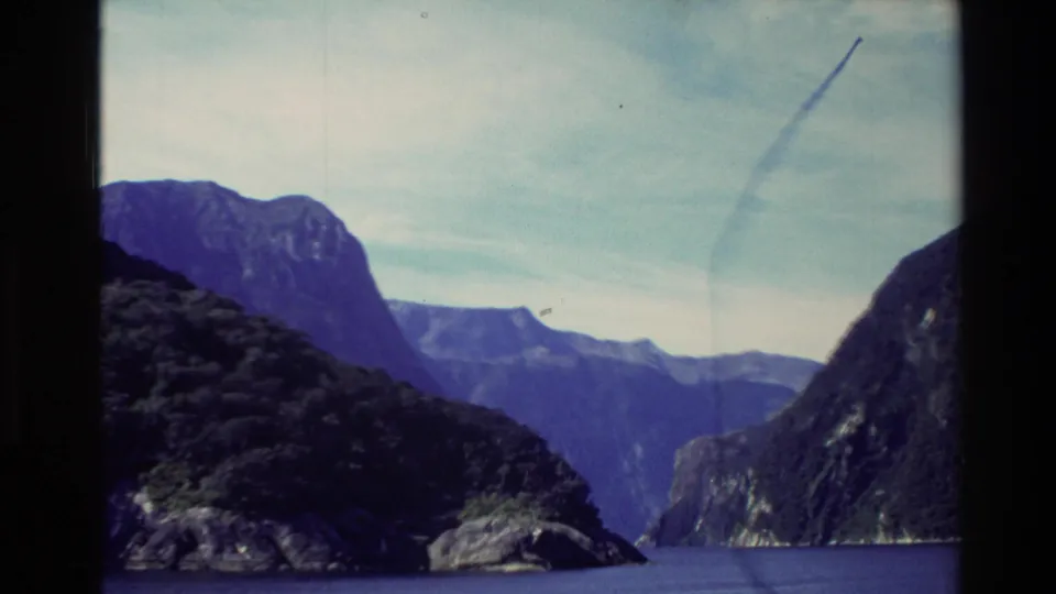 1982 New Zealand cruise view down a fjord, with a dark foreground headland, distant mountain layers, pale sky, and faint contrail.