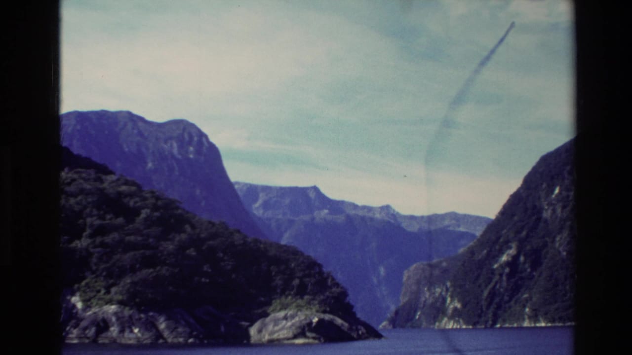 1982 8mm New Zealand travel frame showing a calm lake between forested mountain slopes and distant rocky peaks.