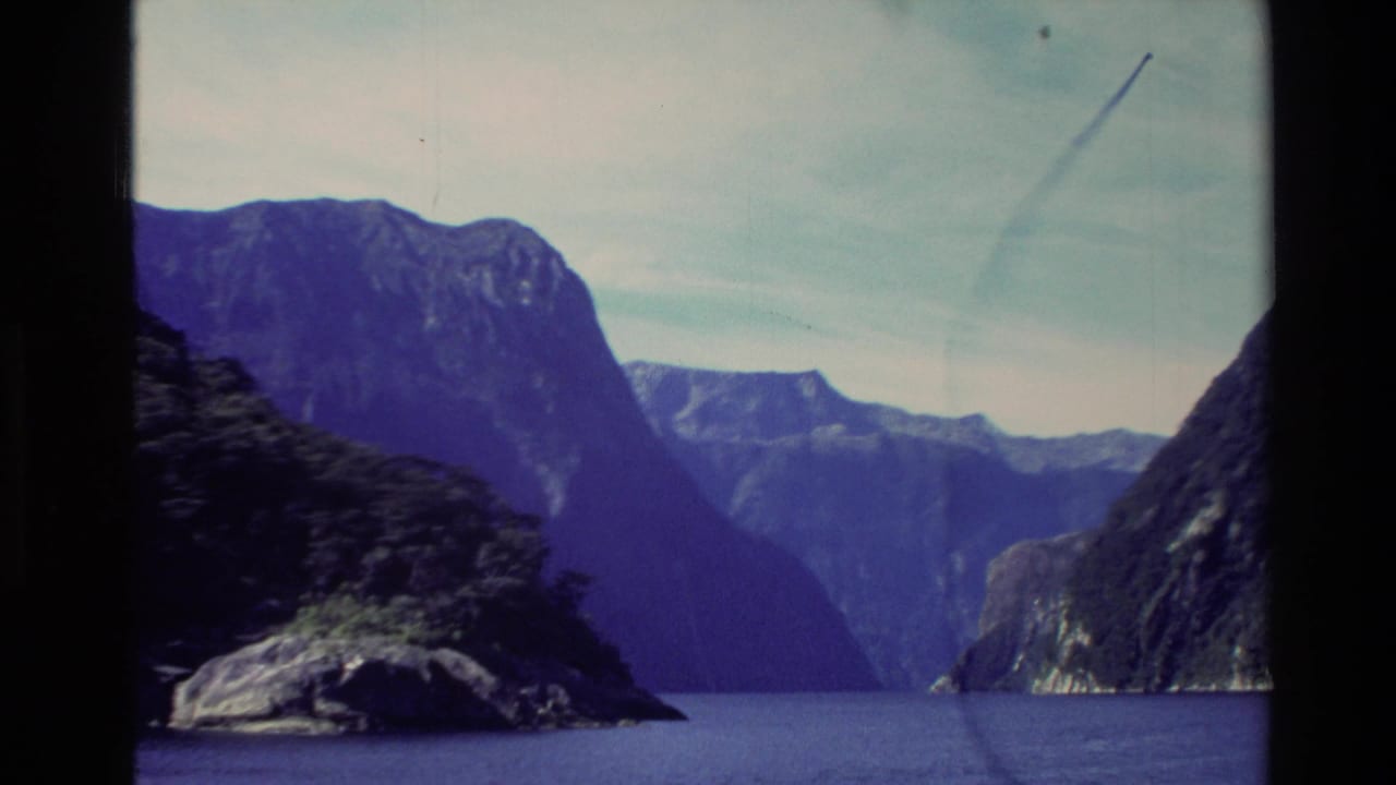 Lake and steep mountains with a dark shoreline in the foreground and pale sky above.