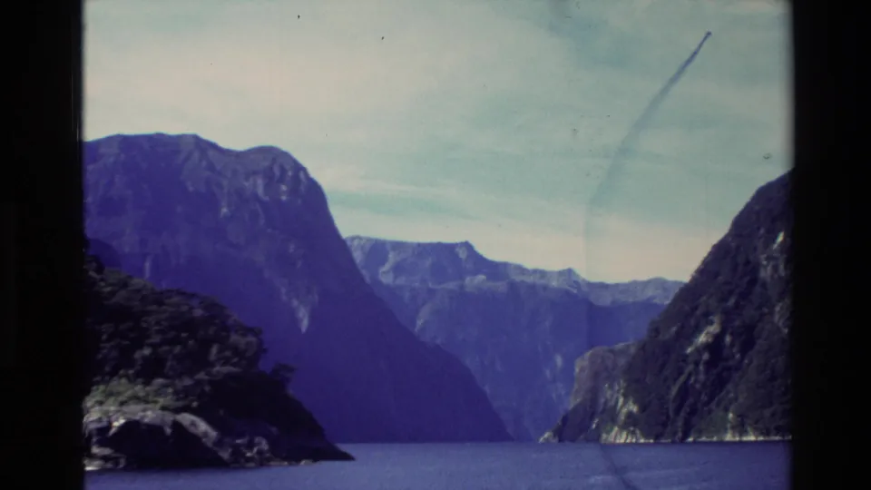 1982 8mm New Zealand frame of a calm lake bordered by shadowed cliffs and layered mountain ridges.