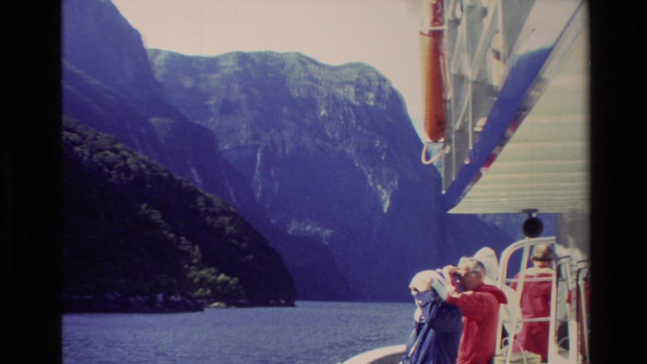 1982 New Zealand 8mm frame showing adults on a boat deck looking across blue water toward steep forested fjord mountains.