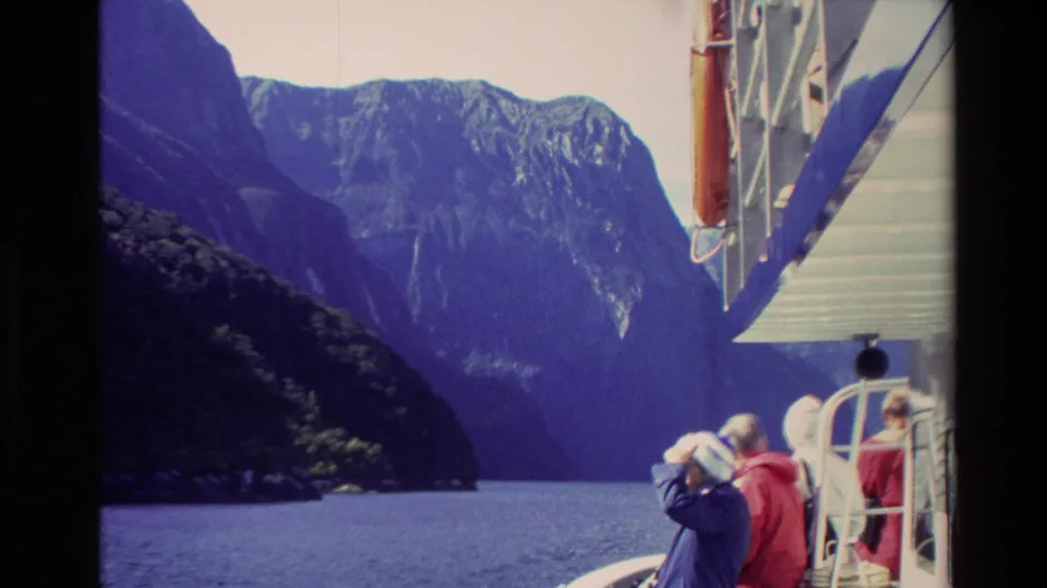 1982 New Zealand home-movie frame with adults in red and blue jackets using binoculars from a boat amid fjord mountains.