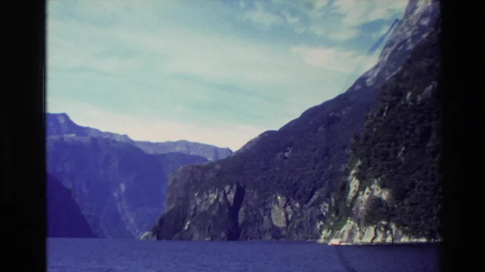 1982 New Zealand 8mm scenic frame looking from water toward steep rocky fjord walls and forested slopes under clouded sky.