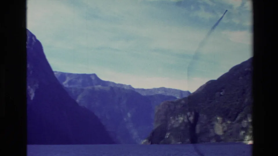 1982 New Zealand 8mm frame with dark foreground mountain slopes, blue water, distant cliffs, and a clouded sky in a fjord landscape.