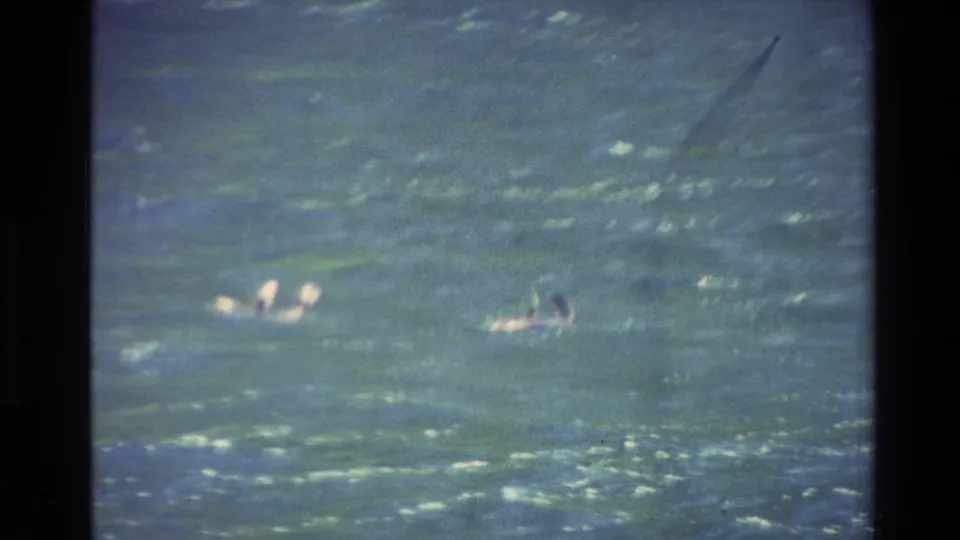 1982 New Zealand home-movie frame showing two distant people near surfboards, partly obscured by rough water and soft focus.
