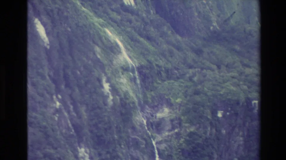 New Zealand 1982 8mm frame of a forested slope dropping toward a lake, with pale rock lines and faded color.