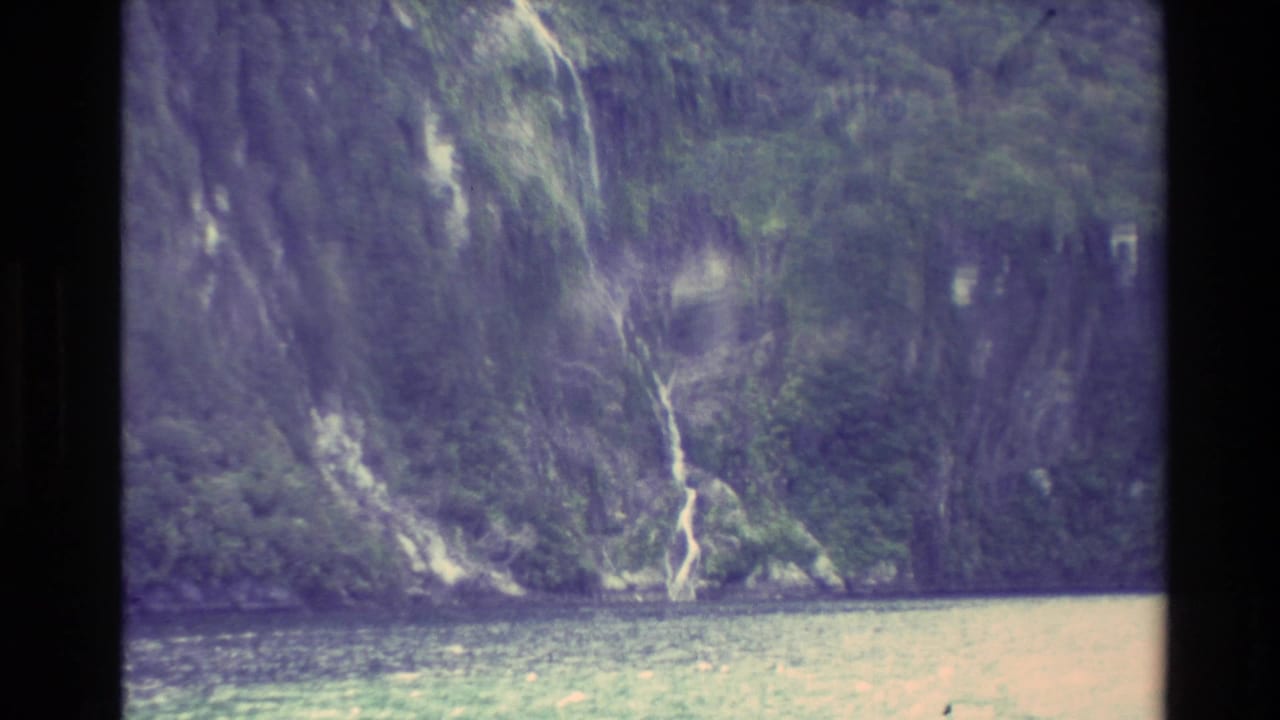 1982 New Zealand 8mm frame showing a steep green and rocky fjord wall with narrow waterfalls and water in the foreground.