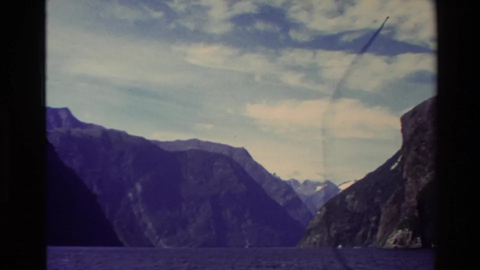 1982 New Zealand 8mm view across deep fjord water toward layered mountain slopes and a partly cloudy sky.