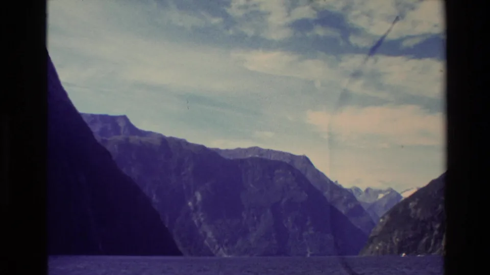 1982 New Zealand home-movie frame looking across fjord water between dark cliffs toward distant mountain forms.