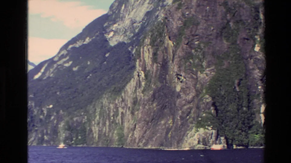 1982 New Zealand 8mm frame of a towering rocky fjord wall, blue water, small waterfalls, and shoreline vegetation.
