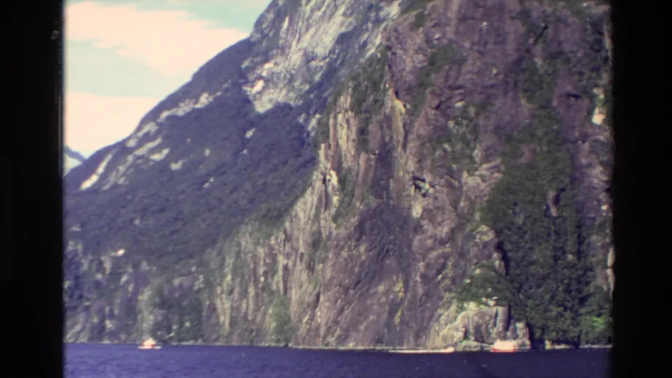 1982 New Zealand 8mm frame of a steep vegetated mountain cliff above blue fjord water, with small boats emphasizing the scale.