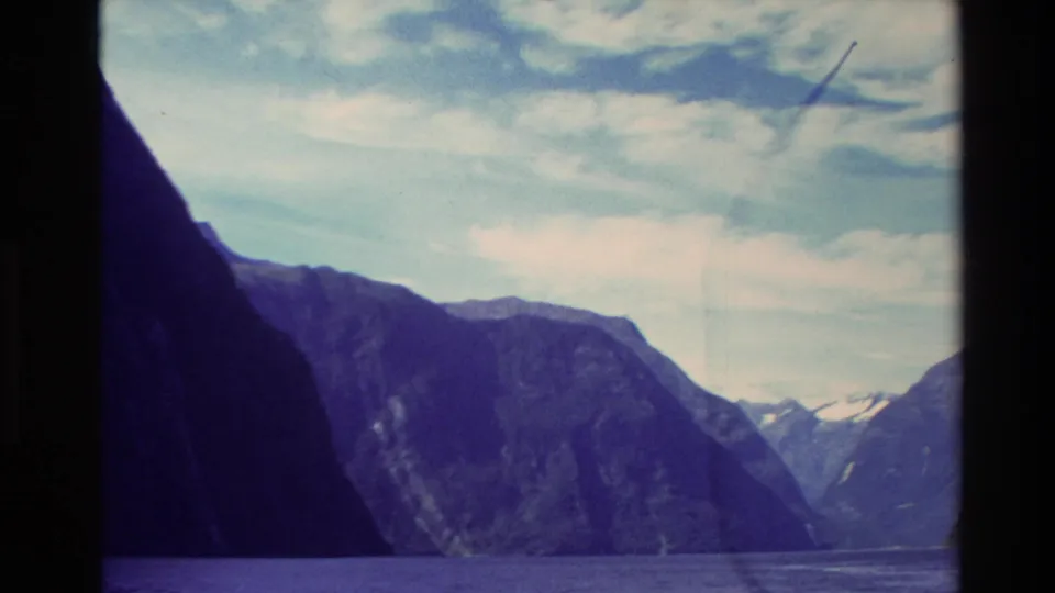 1982 New Zealand 8mm frame showing dark mountain walls around fjord water beneath a pale cloudy sky.