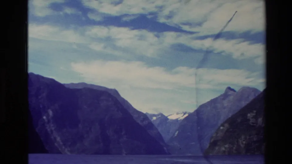 1982 New Zealand 8mm frame of a wide fjord passage with dark mountain slopes, distant snow, and a clouded sky.