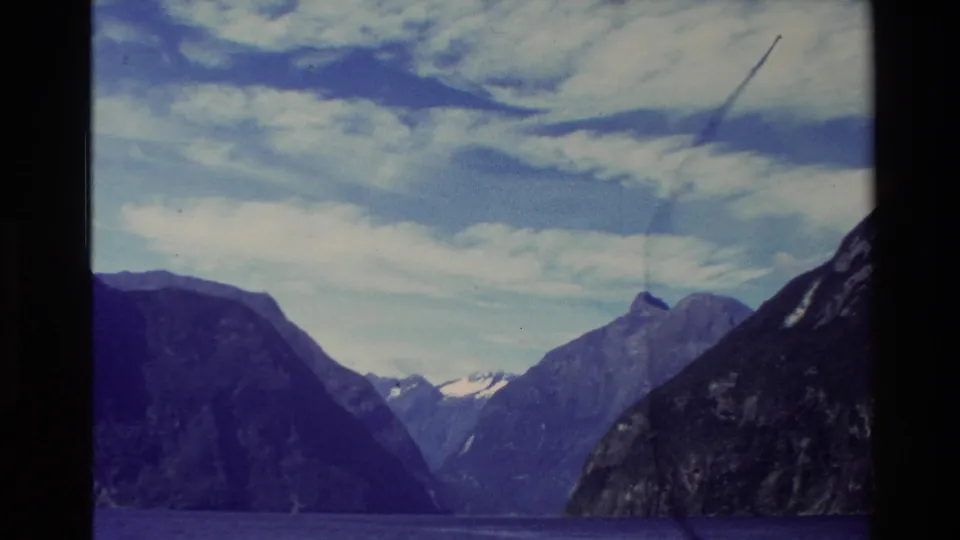 1982 New Zealand 8mm frame looking across fjord water toward steep mountains and distant snow beneath textured clouds.