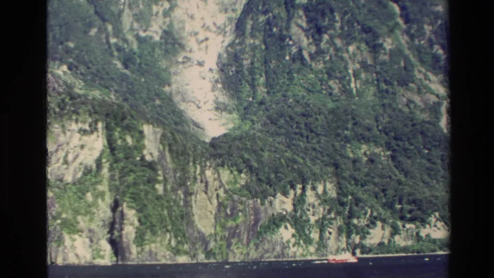 1982 New Zealand aerial frame showing forested cliffs, pale rock, dark water, and a small red boat below.