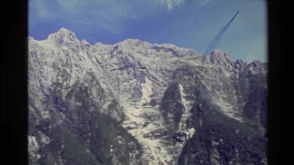 1982 New Zealand travel footage frame with snow-marked mountain ridges, dark rock faces, and a diagonal contrail across the sky.