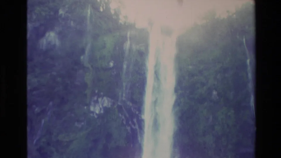 1982 New Zealand 8mm frame showing a tall waterfall dropping through green, shadowed terrain with strong light and soft focus.