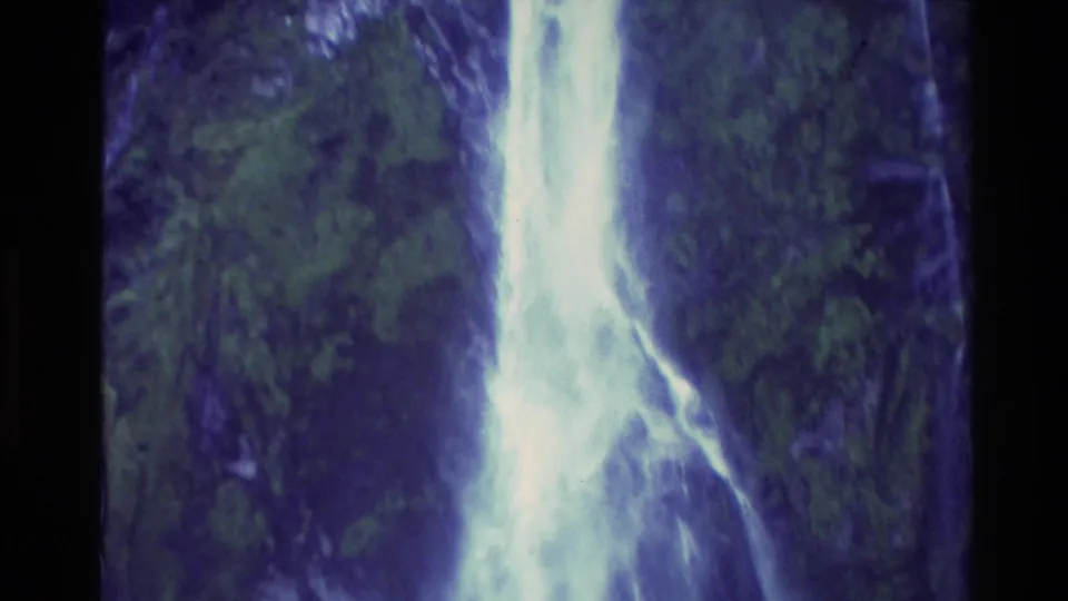1982 New Zealand home-movie frame with a bright vertical waterfall cutting through dark green vegetation and rock.