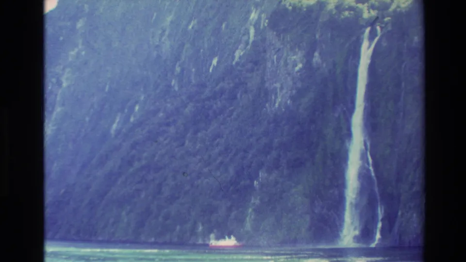 1982 New Zealand 8mm frame showing a tall waterfall on a cliff with a small distant boat on the water for scale.