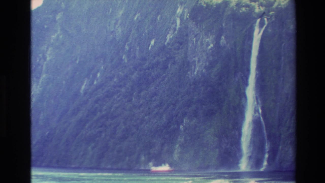 1982 New Zealand 8mm frame of a high waterfall on a dark fjord wall, with a small boat near the base for scale.