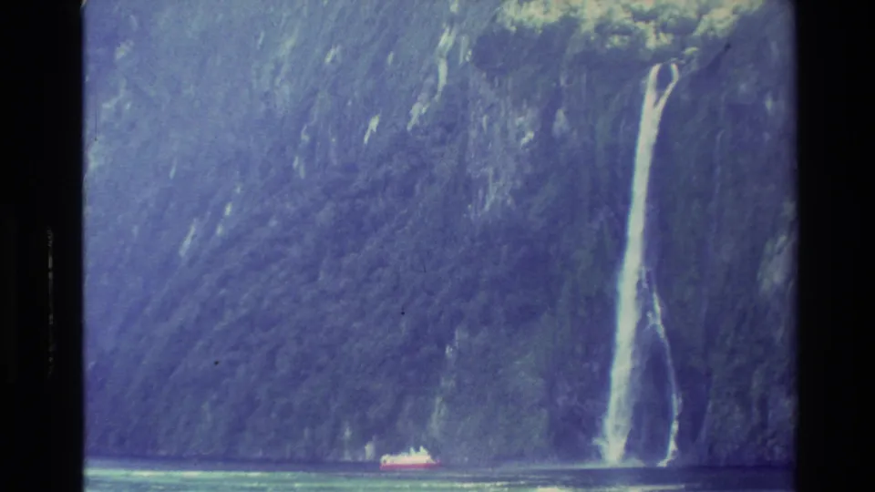 New Zealand fjord scenery from 1982 shows a waterfall pouring down a steep green-blue cliff with a small boat below.