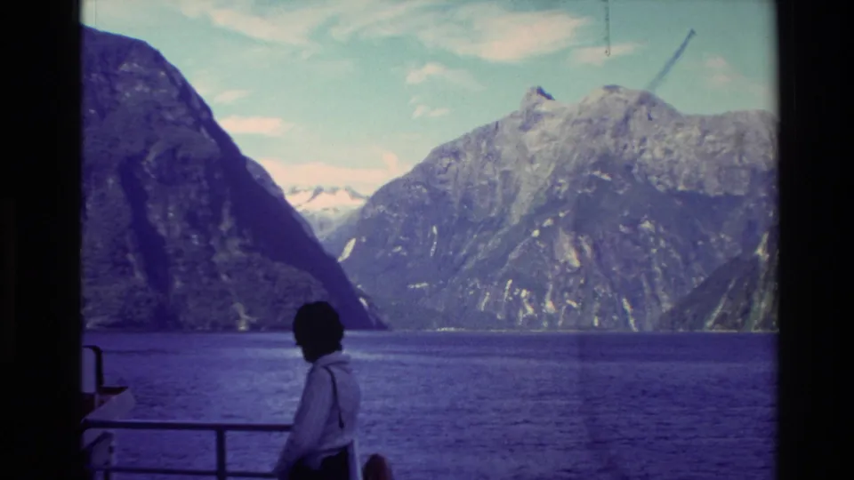 1982 New Zealand home-movie frame with a passenger on a sightseeing boat facing broad fjord water and mountain ridges.