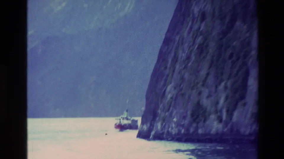 New Zealand 1982 8mm frame of a small boat dwarfed by a steep fjord cliff and hazy mountain backdrop.