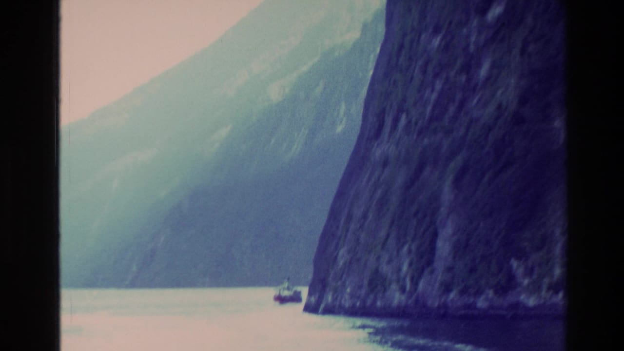 1982 New Zealand 8mm frame showing a small boat dwarfed by dark fjord cliffs and pale mountain slopes across calm water.