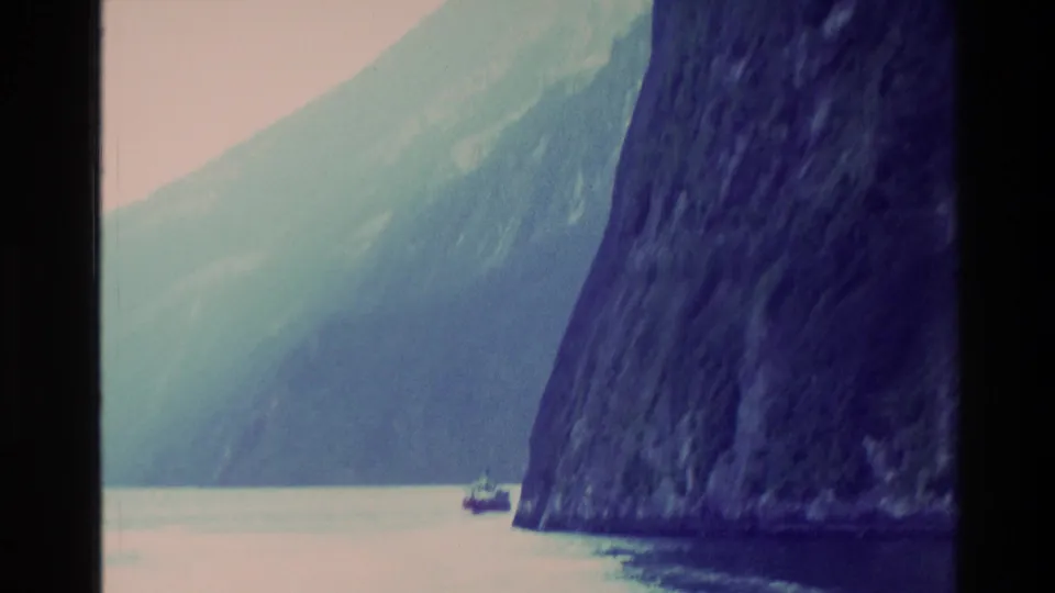 1982 New Zealand 8mm frame showing a small boat dwarfed by dark fjord cliffs and pale mountain slopes across calm water.