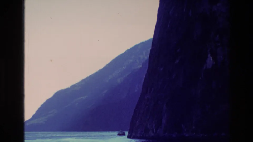 1982 New Zealand 8mm frame of a boat passing near steep fjord cliffs, with layered mountain slopes under a pale sky.