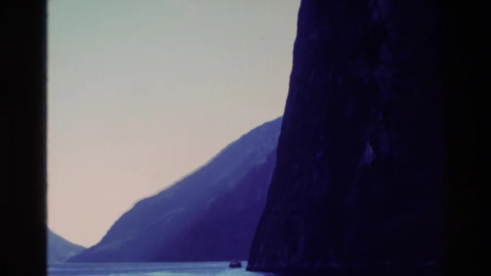 1982 New Zealand 8mm frame dominated by a dark fjord cliff, with calm water and distant mountains visible through faded color.