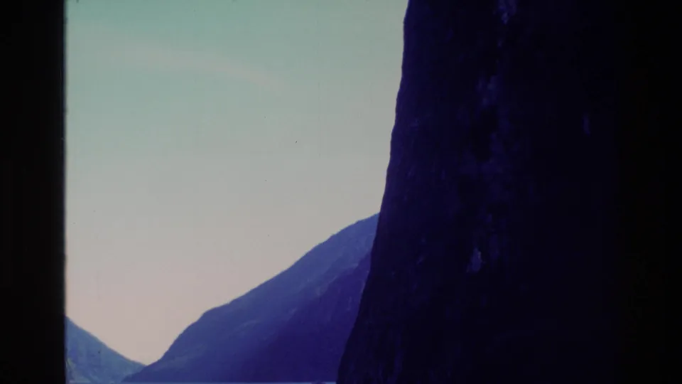 1982 New Zealand 8mm frame showing a steep shadowed fjord wall, distant mountain slope, and calm blue-green water.