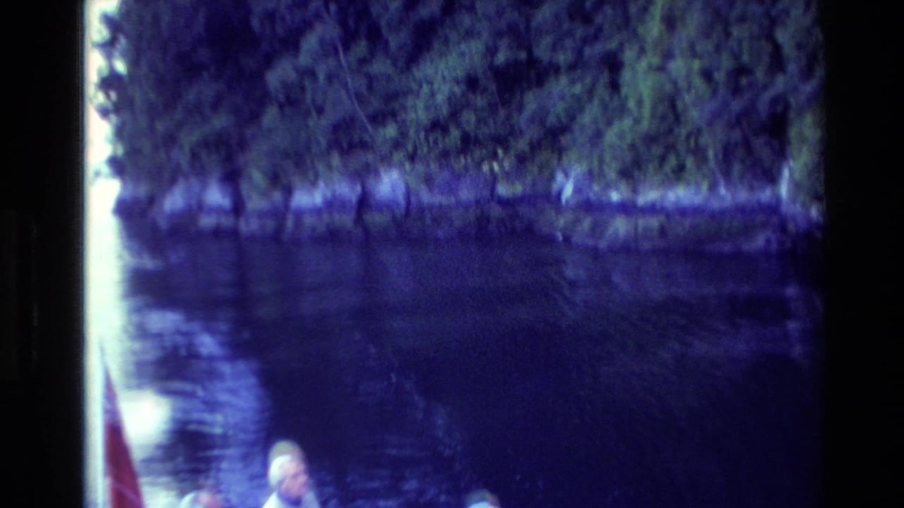 1982 New Zealand home-movie frame with partial boat passengers, calm water, and dense trees along the bank.