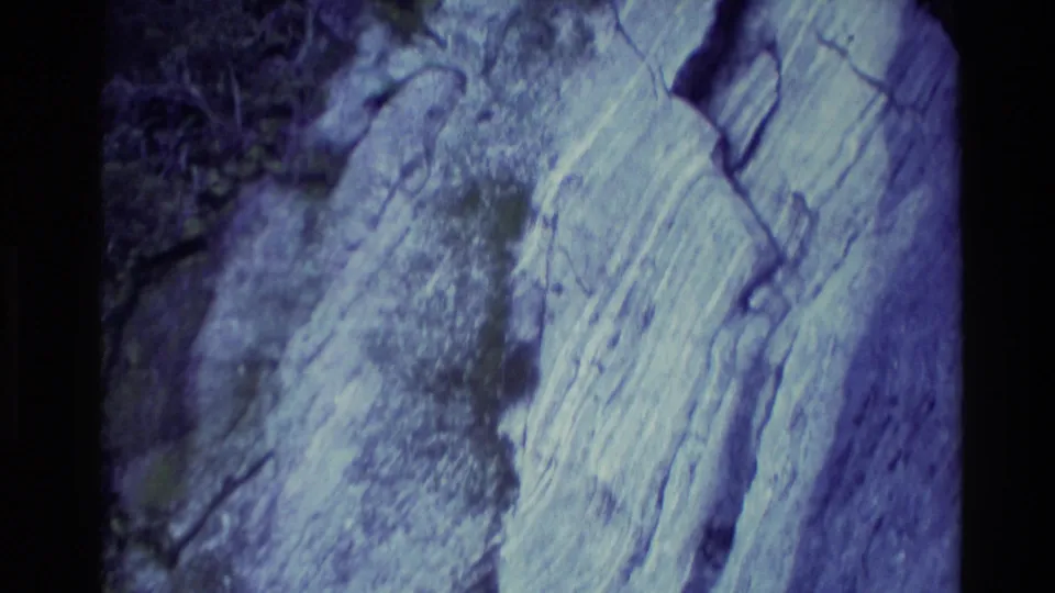 Close 8mm view of a pale New Zealand rock face, showing cracks, vertical streaks, and blue-tinted archival color.