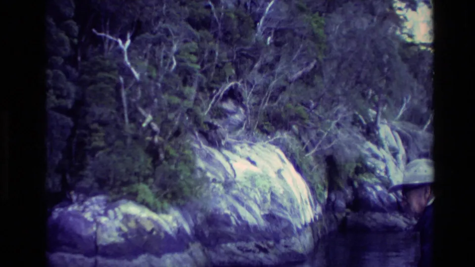 1982 New Zealand home-movie frame showing rocks, bush, a small cascade, and a hat-wearing man partly visible at the lower right edge.