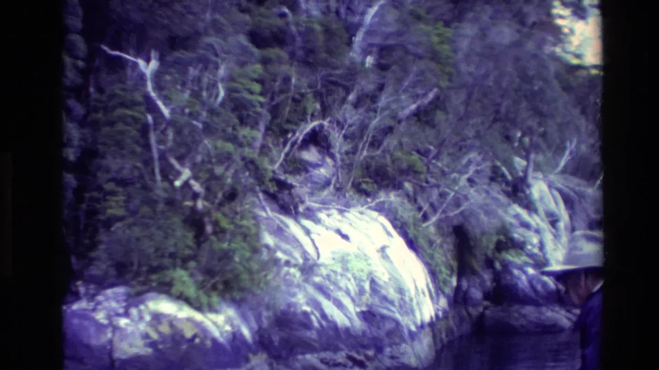 New Zealand 1982 8mm frame with dense bush, pale rock, falling water, and a partly visible man in a broad-brimmed hat at frame edge.