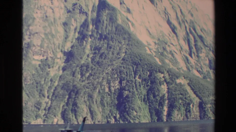 1982 New Zealand 8mm frame of a steep green and rocky mountainside rising above calm water, with part of a boat or ferry visible at the bottom.