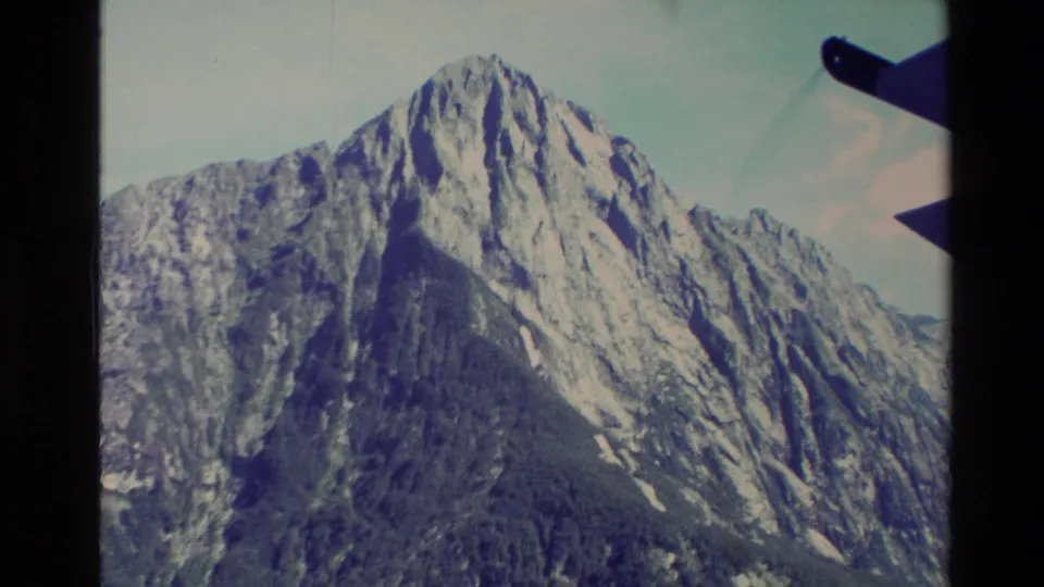 1982 New Zealand 8mm frame of an alpine mountain and ridges viewed from a ferry or boat, with dark vessel hardware visible at the right edge.