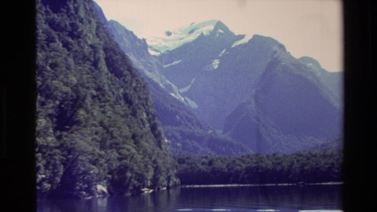 1982 New Zealand 8mm frame showing dark forested slopes, a lake edge, and distant snow-covered mountains under pale sky.