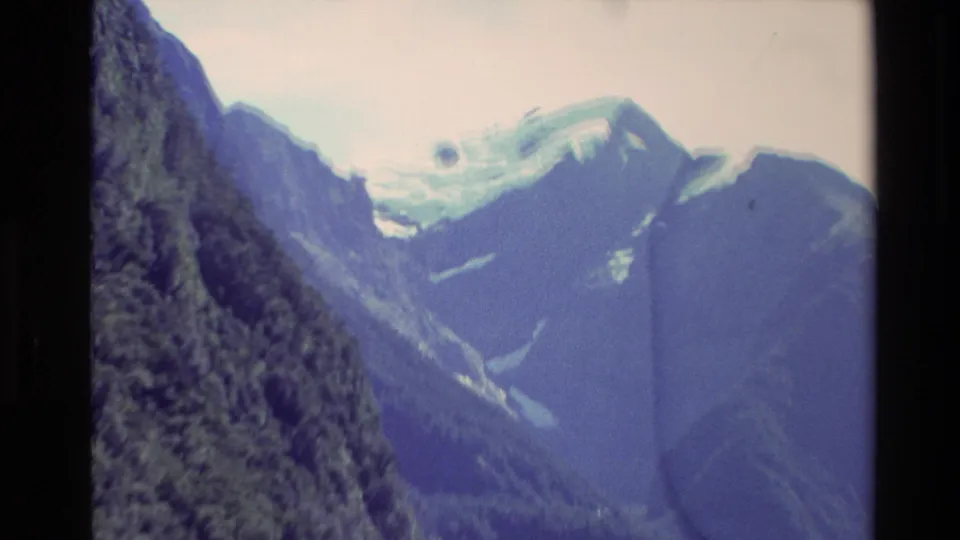 1982 New Zealand home-movie frame with a steep green mountainside, distant snowy ridge, pale sky, and visible 8mm gate edges.