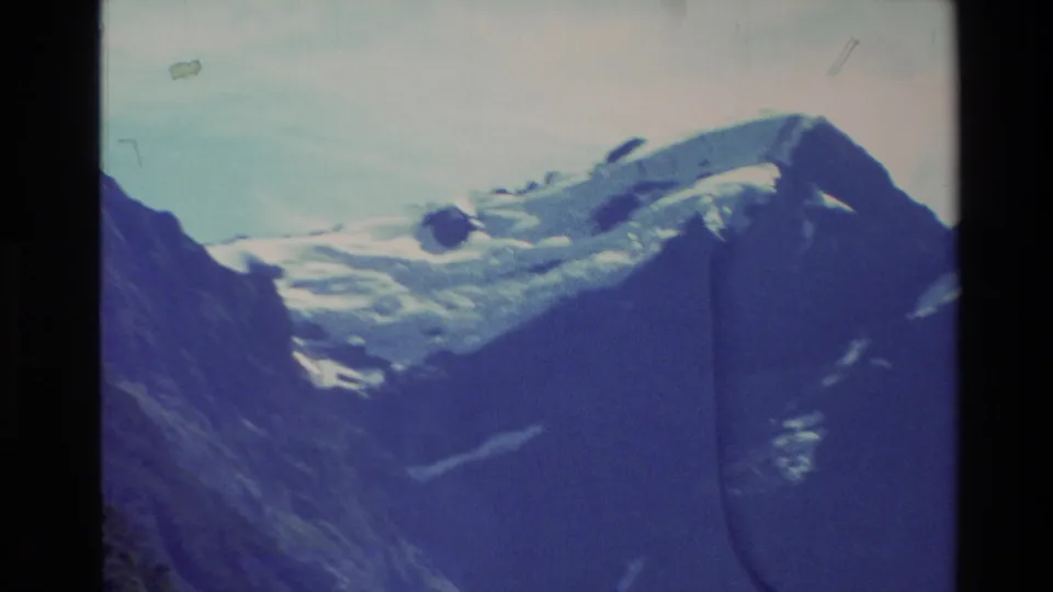 1982 New Zealand home-movie frame showing a snowy mountain ridge, dark adjacent slopes, pale sky, and visible 8mm film artifacts.