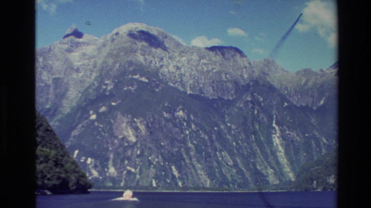 1982 New Zealand 8mm view of steep, snow-streaked mountains above deep blue water, with a small white boat providing scale.