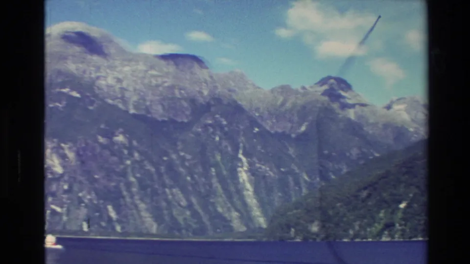 1982 New Zealand home-movie frame showing rugged mountain slopes, patches of snow, clouds, and dark water in a fjord-like setting.