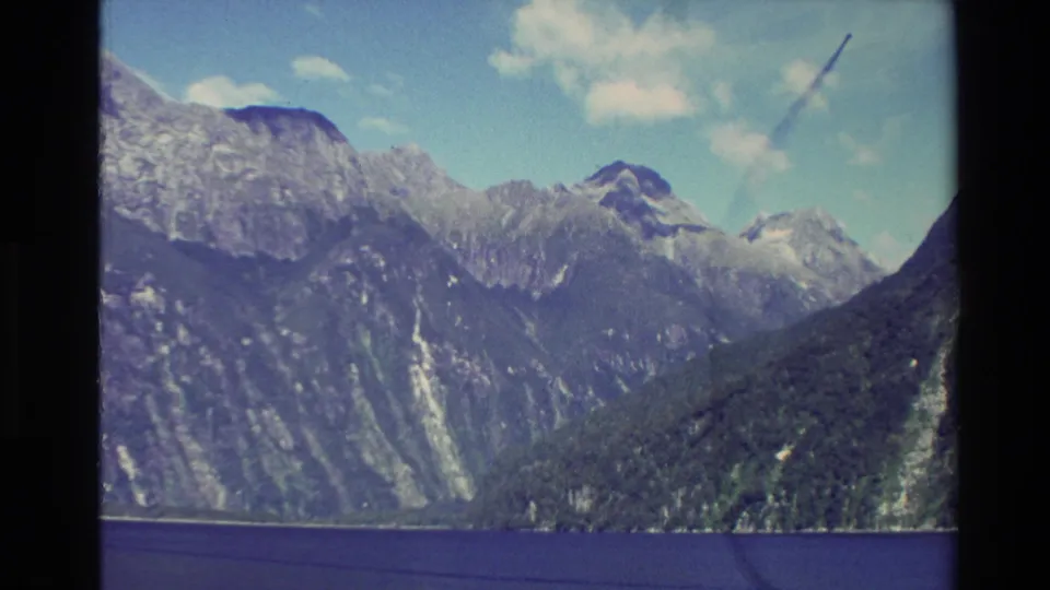 1982 New Zealand 8mm frame with forested slopes, snow-marked peaks, clouds, and a broad foreground of deep blue water.