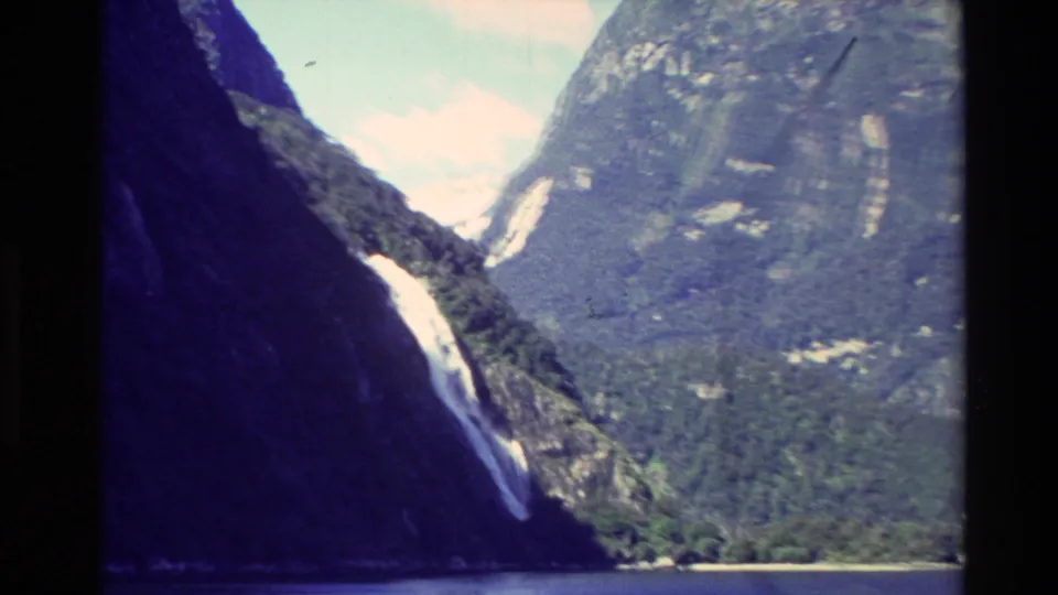 1982 New Zealand 8mm view into a steep mountain valley, with a bright waterfall or snow chute, forested terrain, and water at the base.