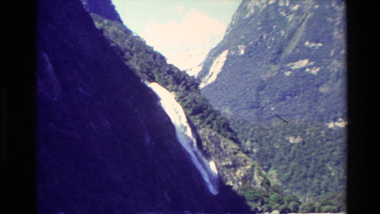 1982 New Zealand 8mm frame showing a waterfall descending through a narrow mountain ravine with forested slopes and bright sky.